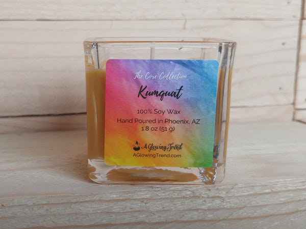 A 1.8 oz glass square containing a yellow-orange Kumquat scented soy candle by A Glowing Trend