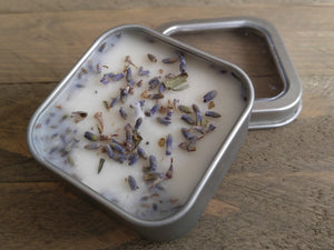 A 1.8 oz square tin containing a white Lavender scented soy candle topped with dried lavender buds by A Glowing Trend.
